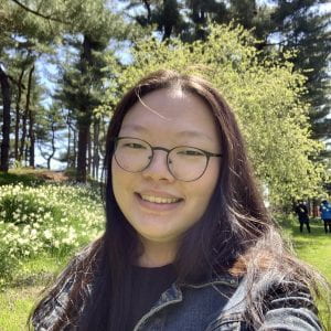 A smiling Asian woman with glasses, dark hair, light skin, and wearing a black long-sleeved shirt is outside on a sunny day.